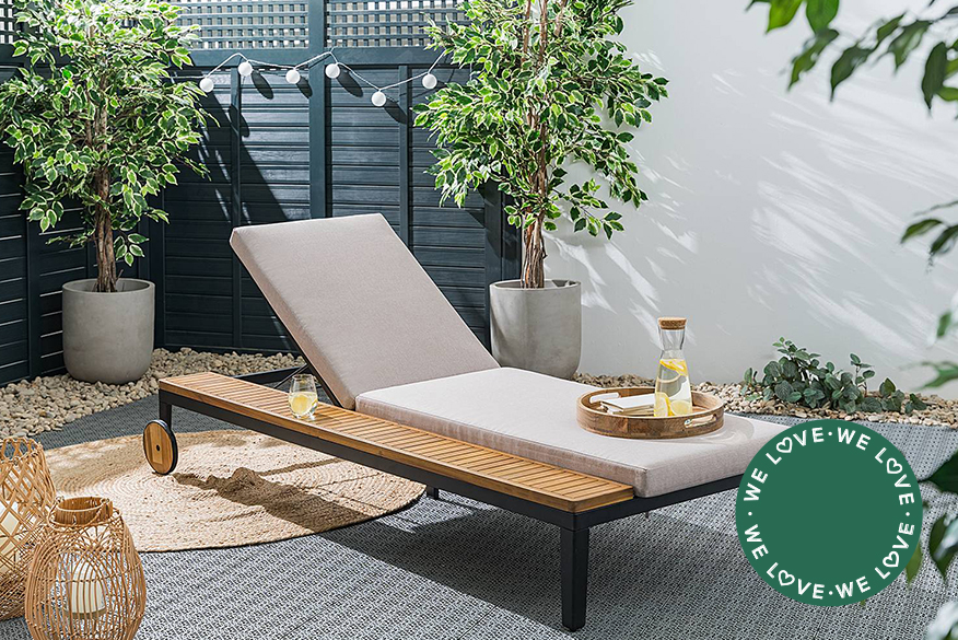 Gartenliege aus Holz und Metall mit grauer Polsterauflage, praktischer Ablagefläche an der Seite und Rollen zum Umstellen je nach Sonnenstand; daneben ein grüner Garten mit einem beigen Sonnenschirm.