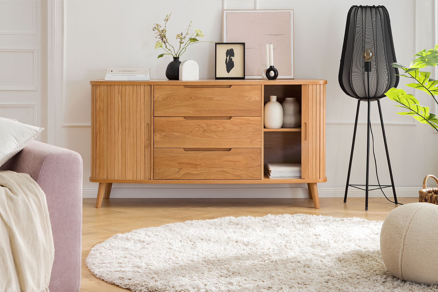 Sideboard Hanck der home24 Exklusivmarke Mørteens aus hellem Eichenholz in Lamellenoptik neben einer Stehlampe aus schwarzem Metall, dazu Deko, ein beiger Teppich und ein beiger Pouf.