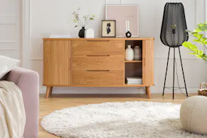 Sideboard Hanck der home24 Exklusivmarke Mørteens aus hellem Eichenholz in Lamellenoptik neben einer Stehlampe aus schwarzem Metall, dazu Deko, ein beiger Teppich und ein beiger Pouf.