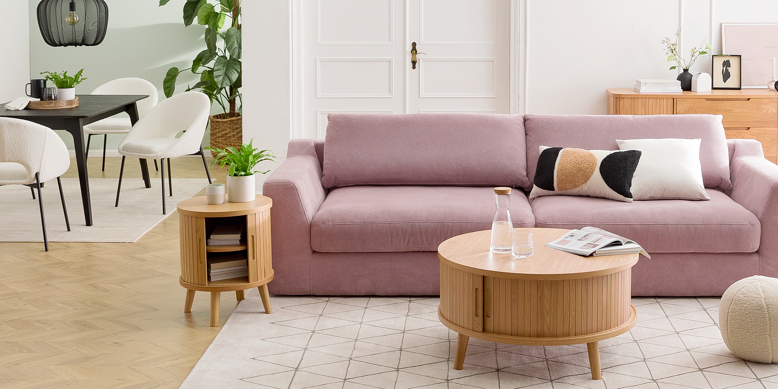 Offener Wohn- und Essbereich mit dem Trend-Bigsofa Dixwell von home24 als Ankerpunkt, dazu Stauraummöbel aus hellem Holz im Retro-Stil sowie ein schwarzer Esstisch, schwarz-weiße Esszimmerstühle, Teppiche, schwarze Lampen und Grünpflanzen.
