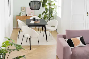 Offener Wohn- und Essbereich mit dem Trend-Bigsofa Dixwell von home24 als Ankerpunkt, dazu Stauraummöbel aus hellem Holz im Retro-Stil sowie ein schwarzer Esstisch, schwarz-weiße Esszimmerstühle, Teppiche, schwarze Lampen und Grünpflanzen.