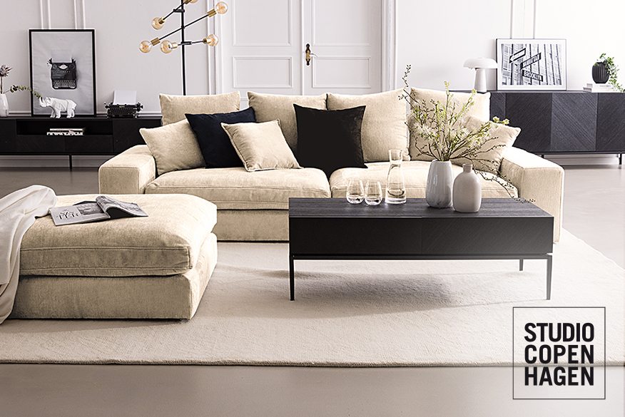 Beiges gemütliches Bigsofa Rodia von Studio Copenhagen mit zusätzlicher Kopfstütze sowie vielen Kissen, passendem Hocker und kombiniert mit Couchtisch, Sideboard und Kommode in elegantem Schwarz.