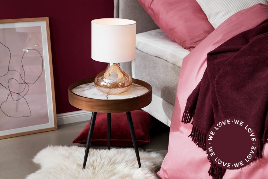 Dunkelbrauner Nachttisch mit schwarzen Beinen und weiß marmorierter Tischoberfläche sowie einer Nachttischleuchte aus lachsfarbenem Glas vor einer beerenfarbenen Wand und neben einem hellgrauen Bett mit Bettwäsche in Beerentönen; daneben ein Detailbild von heimischen Himbeeren, Brombeeren und Blaubeeren.