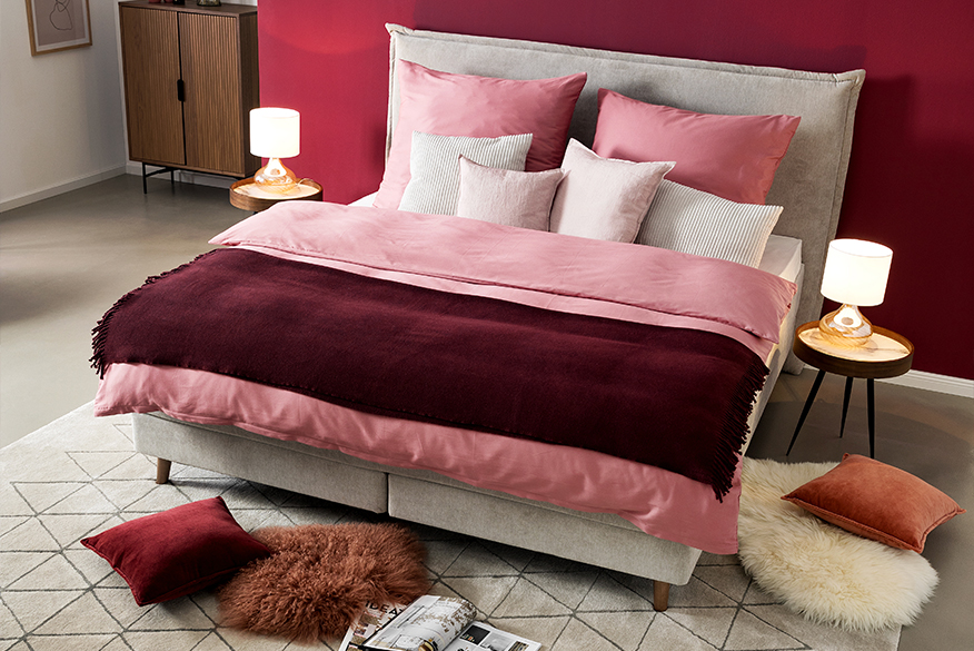Schlafzimmer in roten Berry-Nuancen mit dem Topseller-Bett Avellino der home24 Exklusivmarke Mørteens, bezogen mit Bettwäsche in verschiedenen Rottönen und Weiß, dazu ein grauer Teppich, zwei Nachttische mit Tischleuchten sowie eine Kommode aus dunklem Holz.