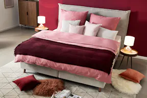 Schlafzimmer in roten Berry-Nuancen mit dem Topseller-Bett Avellino der home24 Exklusivmarke Mørteens, bezogen mit Bettwäsche in verschiedenen Rottönen und Weiß, dazu ein grauer Teppich, zwei Nachttische mit Tischleuchten sowie eine Kommode aus dunklem Holz.