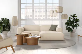 Canapé d'angle Hudson beige et vert foncé dans un salon blanc avec un ensemble de 3 tables basses rondes en bois, des plantes vertes, des lampadaires, des suspensions beiges et un coussin vert, devant une grande baie vitrée.