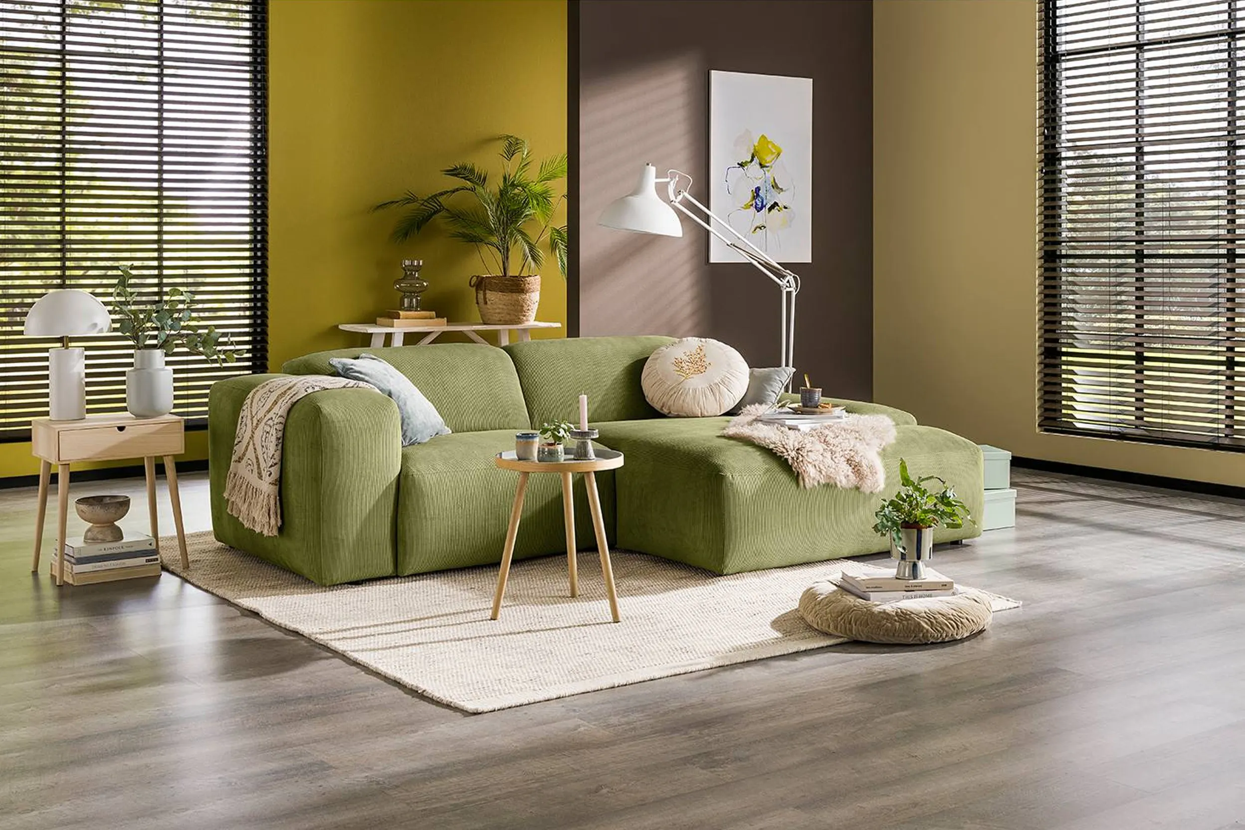 Grünes Sofa, umgeben von beigen Accessoires und kleinen Holzmöbeln, vor einer grün-braunen Wand