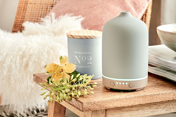 Aroma-Diffuser und Duftkerze von BUTLERS auf einer Holzbank, dekoriert mit Blüten sowie einem Schafsfell im Hintergrund.