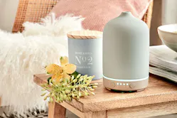 Aroma-Diffuser und Duftkerze von BUTLERS auf einer Holzbank, dekoriert mit Blüten sowie einem Schafsfell im Hintergrund.