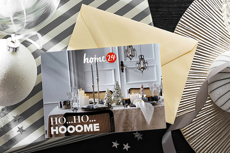 home24 Geschenkgutschein mit Weihnachtsmotiv auf einem schwarzen Tisch mit Weihnachtsgeschenken und -deko in Silber und Weiß