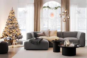 Un salon avec le canapé d'angle Alon en gris clair de la marque exclusive home24, Studio Copenhagen, sur un tapis beige, accompagné d'une table basse ronde noire, d'un pouf gris foncé, de textiles, de suspensions en verre, d'un sapin de Noël décoré, d'étoiles lumineuses et de décorations de Noël.
