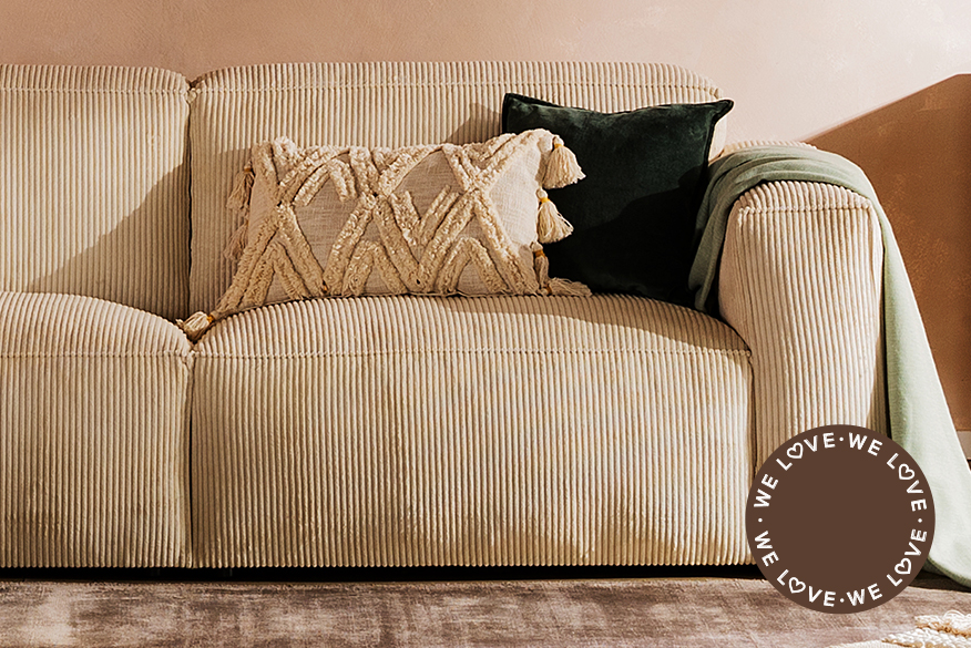 Gemütliches Wohnzimmer im Stilmix aus Boho und Industrial mit einem beigen Cordsofa, einer Holzkommode mit gemusterten Fronten, dazu viele Teppiche, eine coole Stehlampe und Wandregale mit geometrischen Metallrahmen