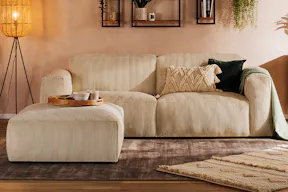 Gemütliches Wohnzimmer im Stilmix aus Boho und Industrial mit einem beigen Cordsofa, einer Holzkommode mit gemusterten Fronten, dazu viele Teppiche, eine coole Stehlampe und Wandregale mit geometrischen Metallrahmen; daneben ein Detailbild einer  Sofaarmlehne aus beigem Cord.