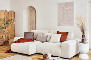 Weißes Ecksofa aus Samt, an einer weißen Wand, mit beigem Teppich, kleinem Couchtisch aus hellem Holz und kleinem weißen Couchtisch, mit einigen Deko-Accessoires.