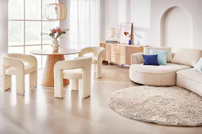 Wohnzimmer mit integrierter Essecke mit einem beigen, abgerundeten Bigsofa auf einem runden, greigen Teppich. Um den runden Holztisch stehen Designer-Esszimmerstühle aus Boucléstoff, ebenfalls in organischer Form; dazu eine Tisch- und Pendelleuchte jeweils aus Rattan.