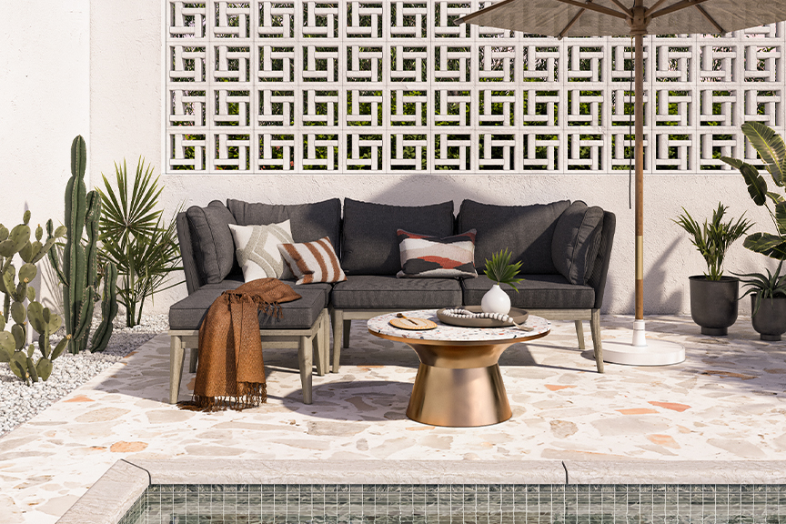 Terrasse im orientalischen Stil mit einem Loungesofa mit grauen Polstern und gemusterten Kissen in Erdtönen, dazu ein kupferfarbener Tisch mit Terrazzo-Platte, ein Sonnenschirm, Kakteen und sonstige Grünpflanzen.