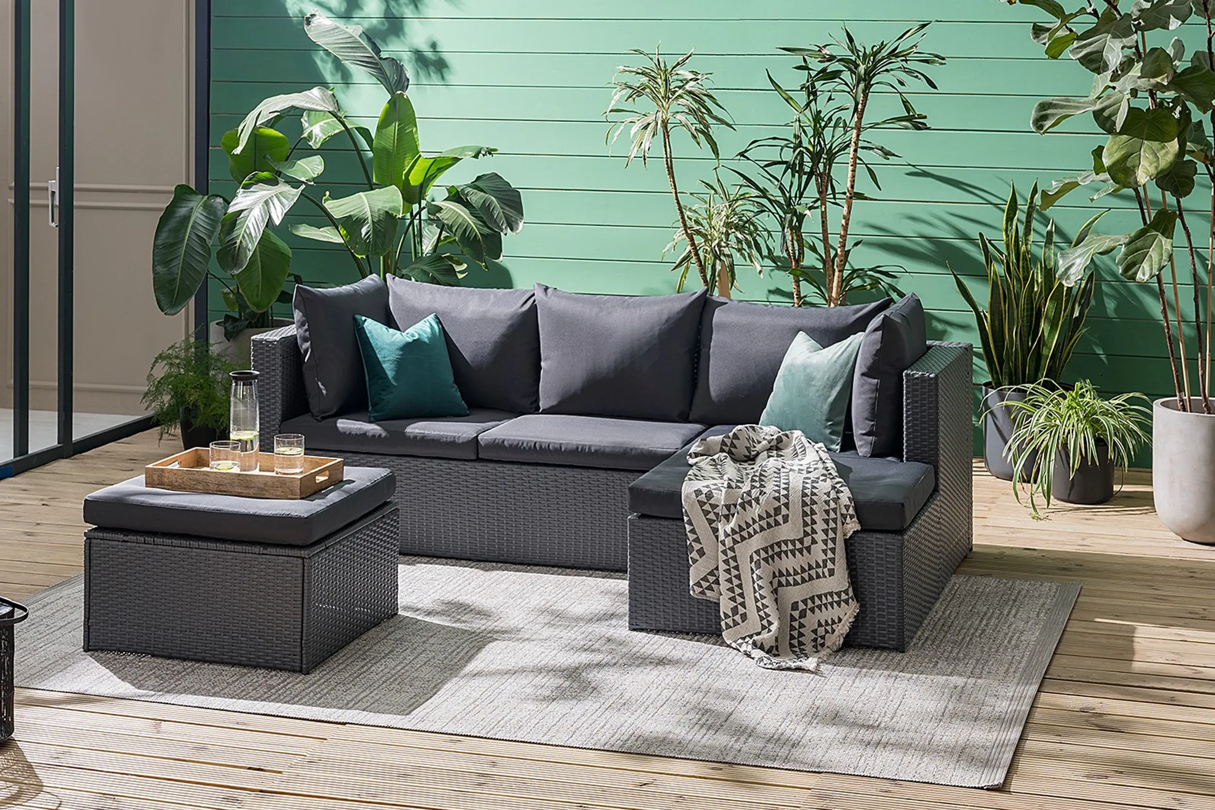 Gartensofa mit Polsterhocker aus Polyrattan in Dunkelgrau vor einer grünen Häuserwand