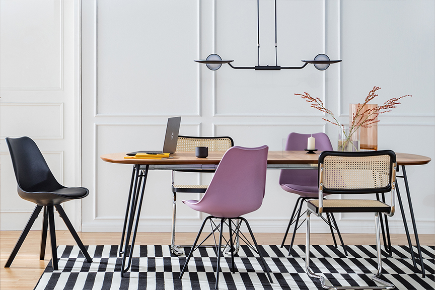 Table de salle à manger avec chaise noire, violette et en cannage sur un tapis graphique noir et blanc, avec suspension en métal et verre et étagère noire.