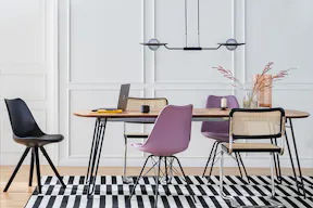 Table de salle à manger avec chaise noire, violette et en cannage sur un tapis graphique noir et blanc, avec suspension en métal et verre et étagère noire.