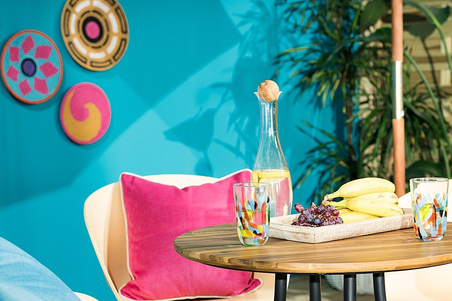 Gartentisch im Industrial-Design mit frischem Obst und einer Wasserkaraffe. Dazu gelbe Schalenstühle mit schwarzen Beinen, bunte Kissen, ein runder Outdoor-Teppich, viele Grünpflanzen in Körben und Hängetöpfen sowie eine Holzleiter.