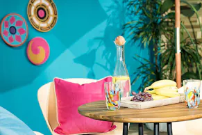 Gartentisch im Industrial-Design mit frischem Obst und einer Wasserkaraffe. Dazu gelbe Schalenstühle mit schwarzen Beinen, bunte Kissen, ein runder Outdoor-Teppich, viele Grünpflanzen in Körben und Hängetöpfen sowie eine Holzleiter.