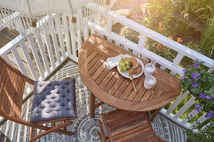 Kleiner Balkon mit praktischen Klappmöbeln von BUTLERS aus dunklem Holz, mit blauem Sitzkissen und weißem Frühstücksgeschirr. 
