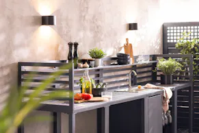 Schwarzes Gartenküchenset von home24 mit verschiedenen Ablageflächen, auf denen Holzbretter, Tomaten, Olivenöl, Kräuter, schwarzes Geschirr und mehr Grillzubehör Platz finden.