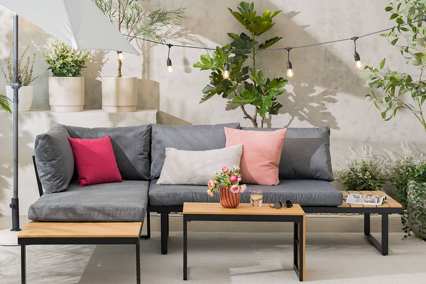 Gartenloungeset Marinella der home24 Exklusivmarke Jardi im Industrial-Stil mit grauen Polstern und bunten Kissen, dazu ein hellgrauer Sonnenschirm, eine Lichterkette und viele Blumenkübel mit Grünpflanzen.