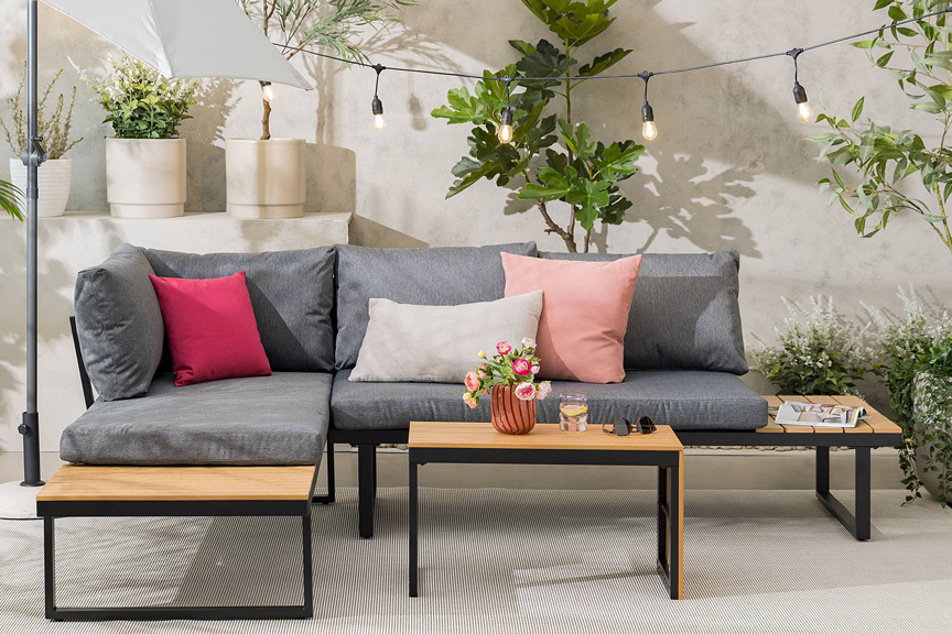 Gartenloungeset Marinella der home24 Exklusivmarke Jardi im Industrial-Stil mit grauen Polstern und bunten Kissen, dazu ein hellgrauer Sonnenschirm, eine Lichterkette und viele Blumenkübel mit Grünpflanzen.