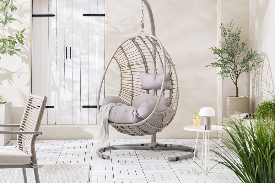 Hängesessel Kayoa von home24 Outdoormarke Jardi aus Polyratten mit bauschigen, grauen Polstern, daneben ein Beistelltisch mit einem Glas Limonade und einer kleinen Outdoorlampe.