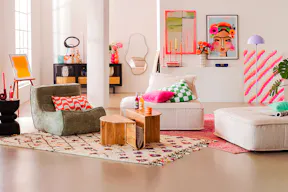 Buntes Boho-Esszimmer in knalligen Neonfarben: Ein bequemer Sessel aus grünem Boucléstoff sowie ein Sessel mit passendem Polsterhocker aus beigem Cord, dazu ein Couchtisch aus Holz, der praktischen Stauraum für Zeitschriften bietet, während ein schwarzes Sideboard mit Wiener Geflecht Retro-Charme ins Spiel bringt. Kunterbunte Wandbilder und Textilien sorgen für farbenfrohe Boho-Stimmung.