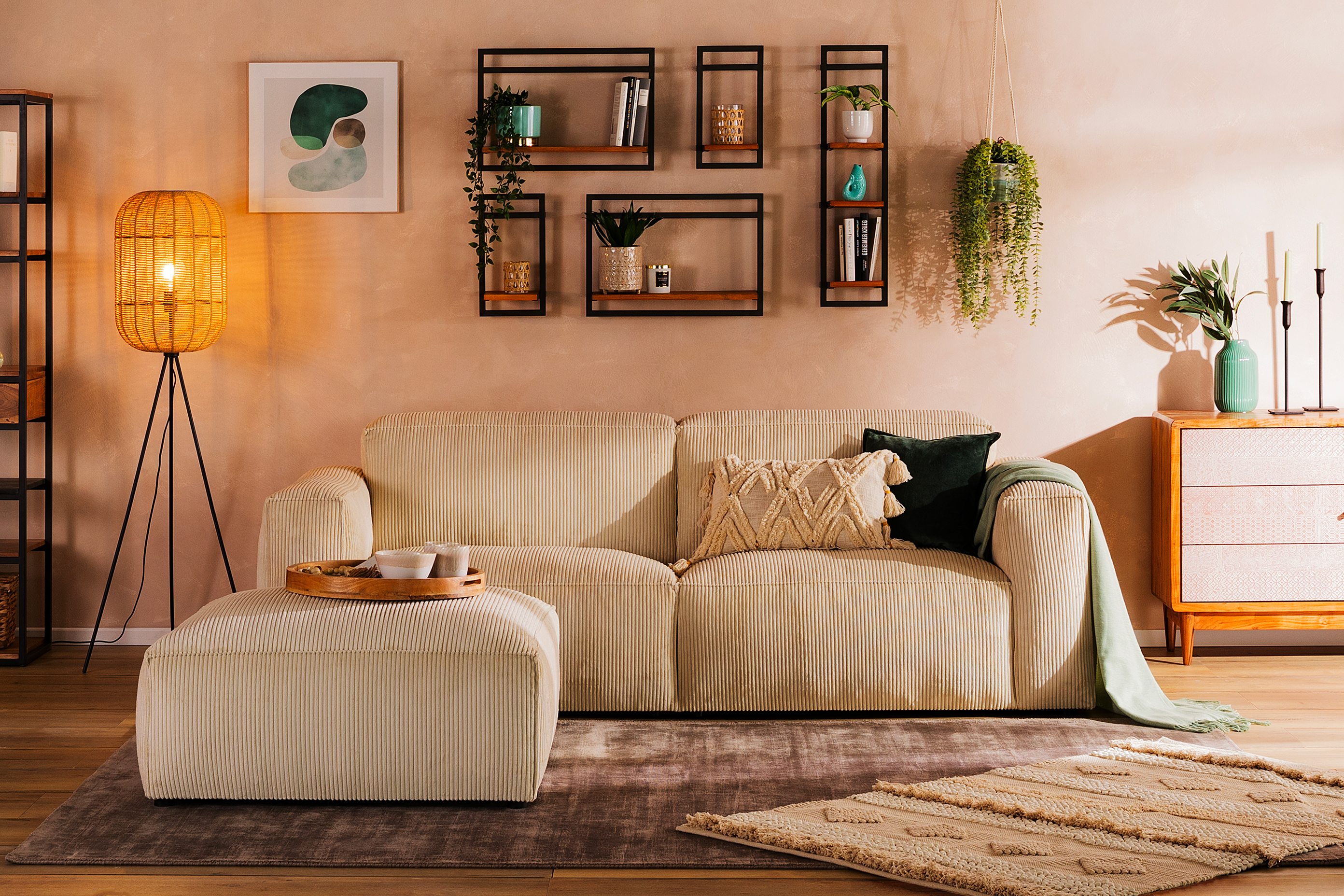 Gemütliches Boho-Wohnzimmer mit einem beigen Cordsofa, einer Holzkommode mit gemusterten Fronten, dazu viele Teppiche, Grünpflanzen, eine coole Stehlampe und Wandregale mit geometrischen Metallrahmen.