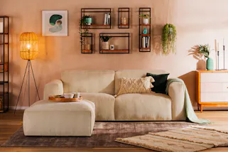 Gemütliches Boho-Wohnzimmer mit einem beigen Cordsofa, einer Holzkommode mit gemusterten Fronten, dazu viele Teppiche, Grünpflanzen, eine coole Stehlampe und Wandregale mit geometrischen Metallrahmen.