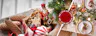 Traditionelle Weihnachtsanhänger wie rote Kugeln, Plätzchenanhänger und ein roter Nussknacker; daneben eine klassisch gedeckter Tisch mit Tannengrün, roten Farbdetails und goldenem Besteck.