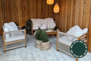 Jardin aménagé de façon chaleureuse pour la période hivernale, avec des meubles d’extérieur en corde, des guirlandes lumineuses, des textiles douillets, des branches de sapin et des bougies.