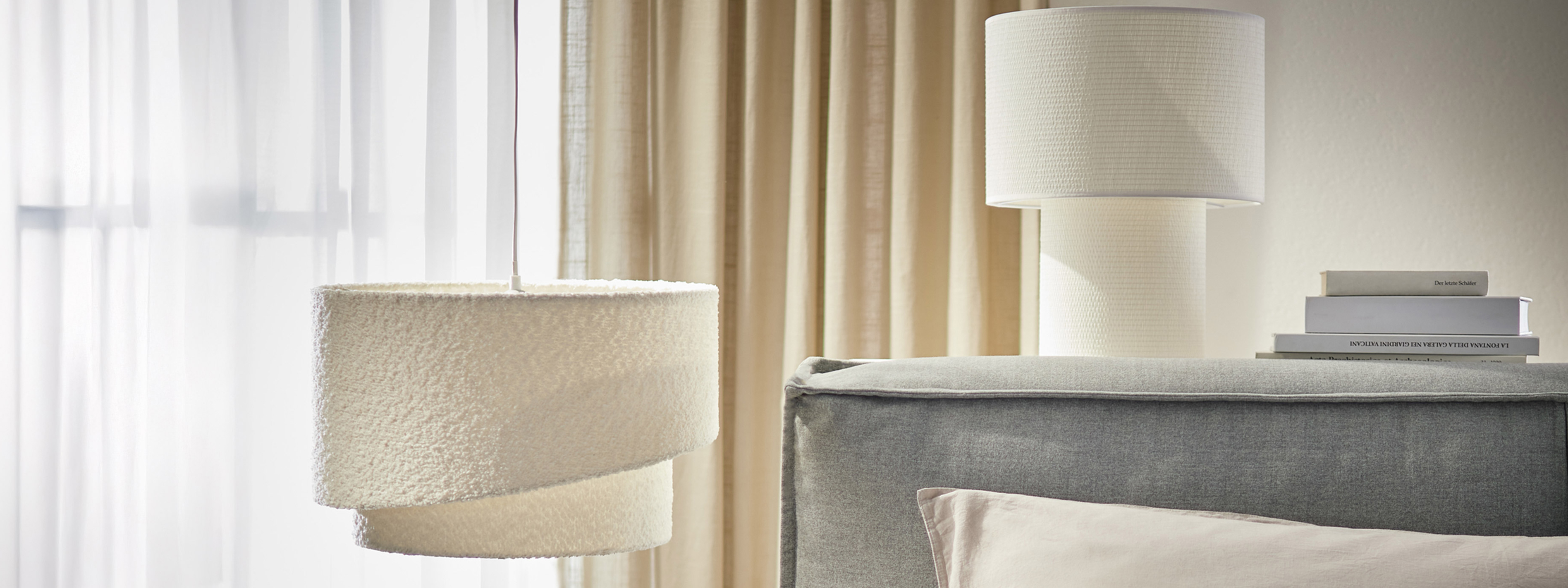 Beige-weiße Pendelleuchte aus Bouclé sowie eine weiße Tischleuchte aus Stoff, dazu Boxspringbett KINX in Grau. 