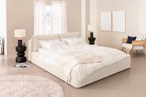 Minimalistisch eingerichtetes Schlafzimmer mit einem weißen Bett in voluminösem, cremeweißem Cordstoff und vielen luftigen Textilien; daneben ein Bild von einem Himmel mit weißen Wölkchen.