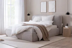 Erholsames Schlafzimmer in warmen Grau-, Beige- und Holztönen mit einem grauen Polsterbett, einem dunkelgrauen Sessel, passender Bettwäsche und einem Sideboard aus hellem Holz.