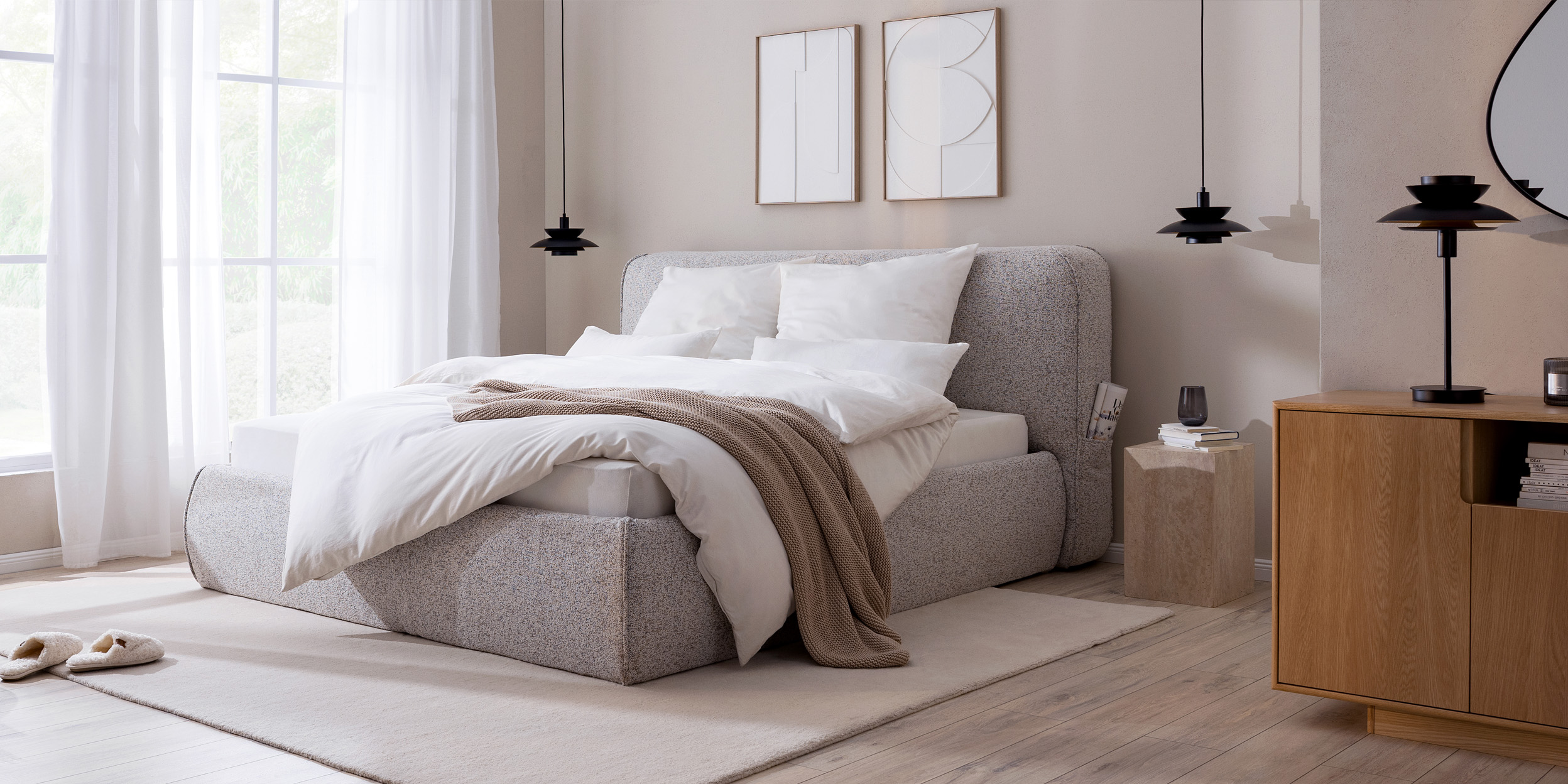 Erholsames Schlafzimmer in warmen Grau-, Beige- und Holztönen mit einem grauen Polsterbett, einem dunkelgrauen Sessel, passender Bettwäsche und einem Sideboard aus hellem Holz.