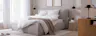 Erholsames Schlafzimmer in warmen Grau-, Beige- und Holztönen mit einem grauen Polsterbett, einem dunkelgrauen Sessel, passender Bettwäsche und einem Sideboard aus hellem Holz.