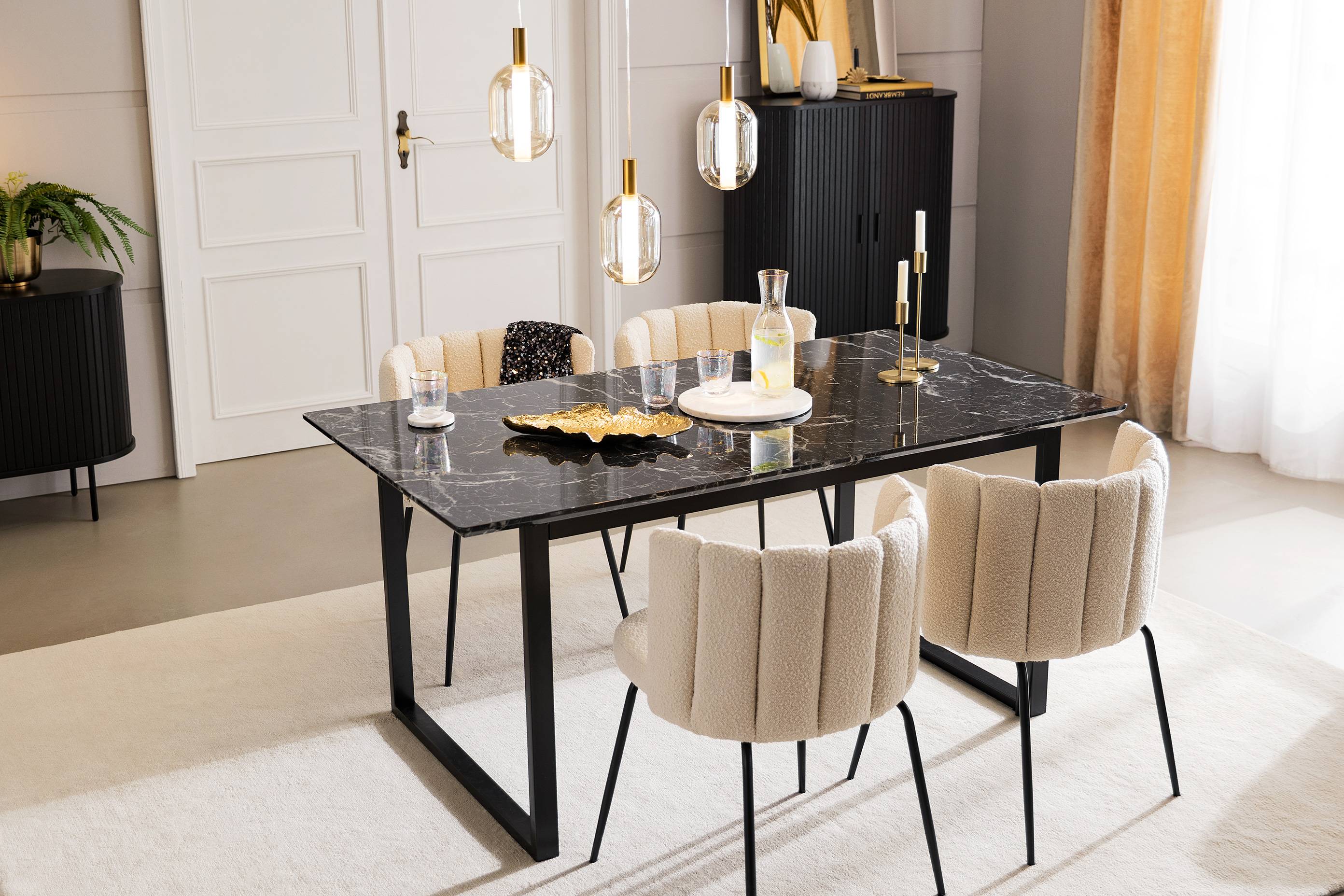 Eleganter Essbereich im Glamourstil mit eleganten Stühlen in beigem Boucléstoff und einem Tisch in Marmoroptik, darauf goldene Schalen, ein Marmortablett und Gläser mit Goldrand, im Hintergrund schwarze Stauraummöbel mit edlen Lamellenfronten.