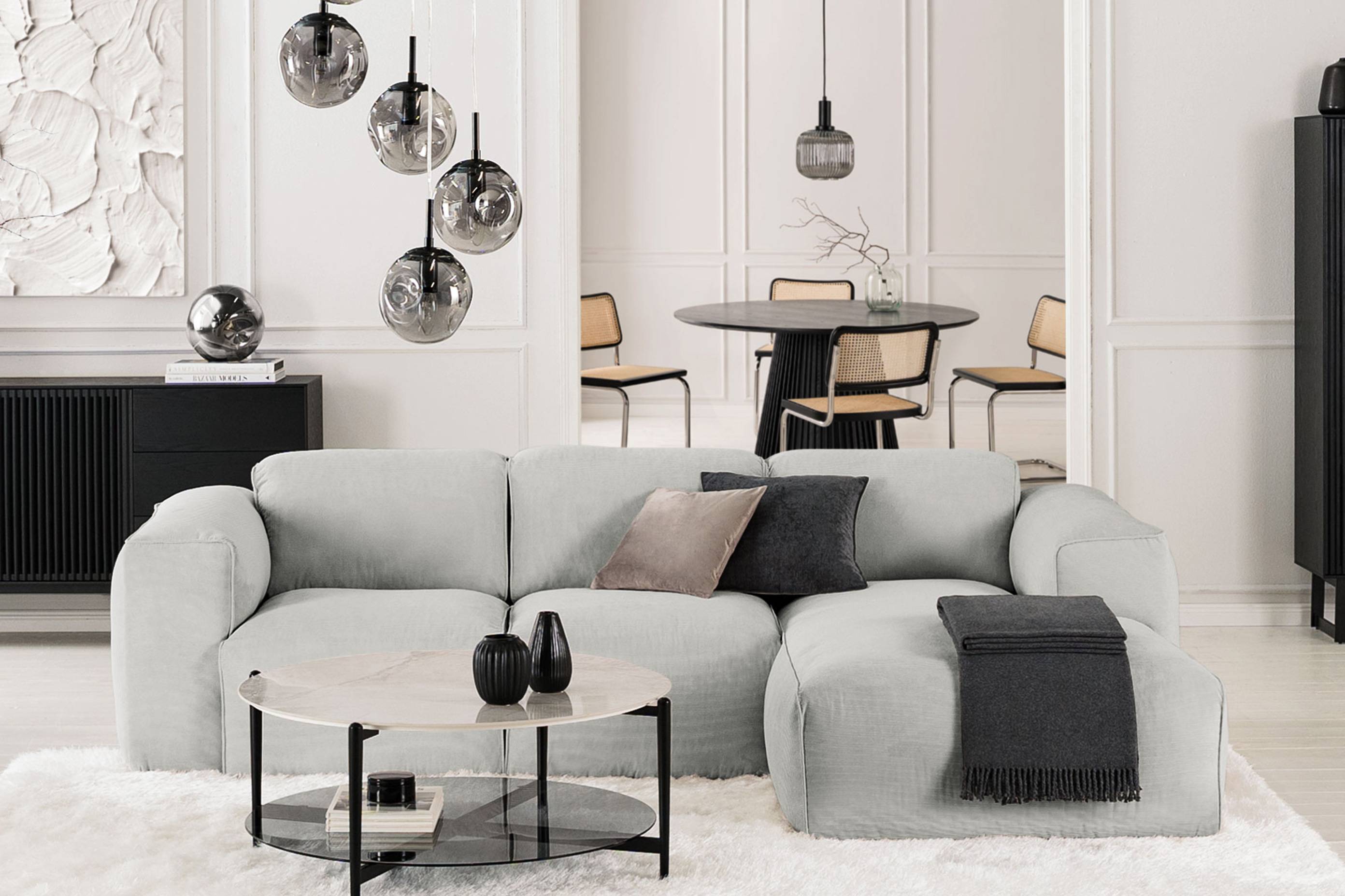 Modern eingerichtetes Wohnzimmer in unifarben Weiß sowie Grau und Schwarz, Sofa Hudson in der Mitte des Raumes, dazu skulpturale Leuchten aus Rauchglas, die schwarzen Stauraummöbel in Lamellen-Optik stammen aus der Serie Qara.