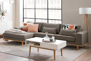 Wohnzimmer eingerichtet im gemütlichen Skandi-Wohnstil mit einem grauen Ecksofa, einem beigen Teppich sowie weiteren Möbeln im skandinavischen Holz-Weiß-Mix von der home24 Exklusivmarke Mørteens, dazu Dekoration mit Kerzen, Textilien, Vasen und vielen verspielten Details.