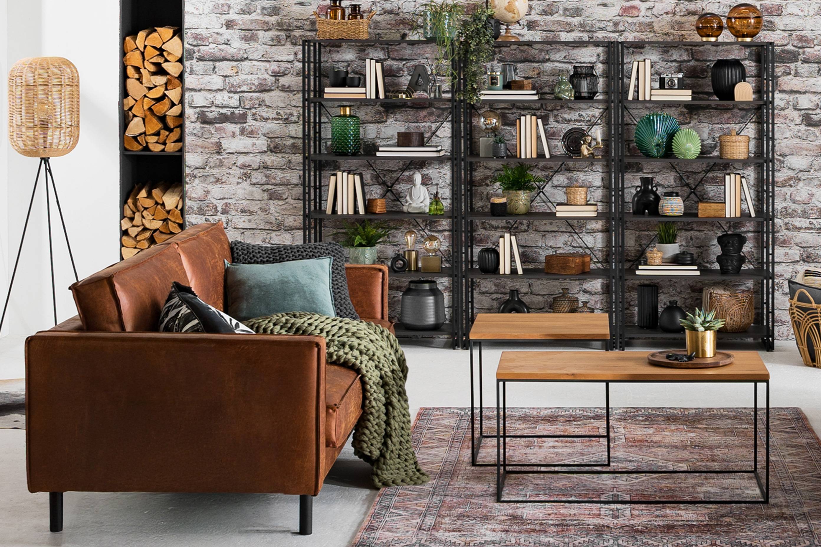 Wohnzimmer im Industrial-Stil mit braunen Ledersofas, zwei Couchtischen im coolen Holz-Metall-Mix und einem offenem Metallregal vor einer Backsteinwand.