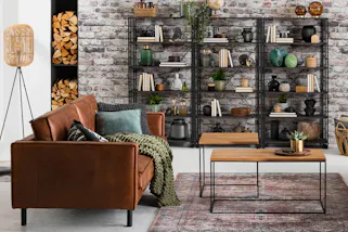 Wohnzimmer im Industrial-Stil mit braunen Ledersofas, zwei Couchtischen im coolen Holz-Metall-Mix und einem offenem Metallregal vor einer Backsteinwand.