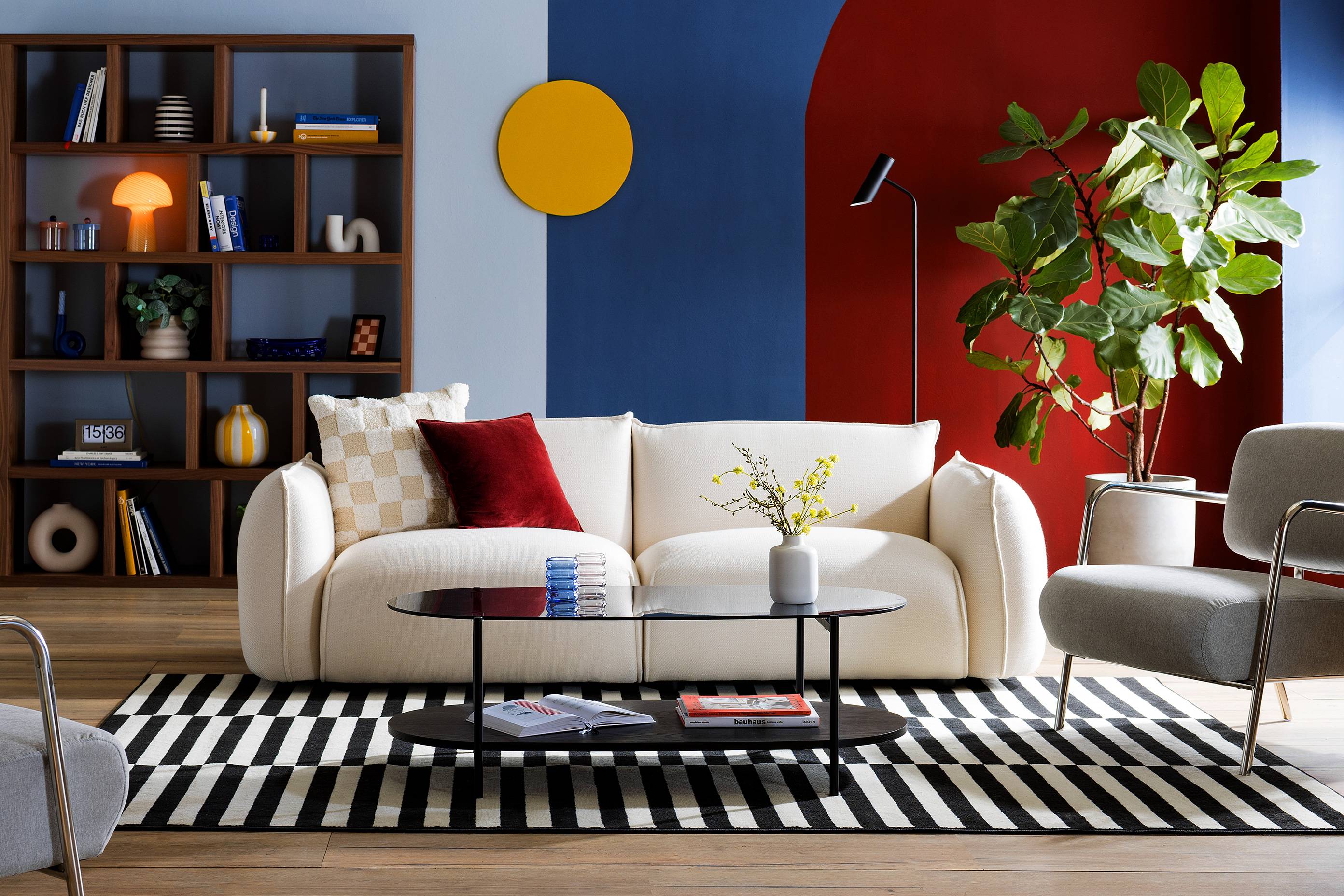 Wohnzimmer in blau-roter Farbgestaltung, inspiriert von den Designepochen Bauhaus und Mid-Century, im Zentrum ein helles Sofa, dazu graue Sessel mit Chromgestell und ein Couchtisch aus Rauchglas, dahinter ein großes Regal mit dunklem Walnussfurnier; dekoriert ist das Setting mit grafisch gemusterten Textilien und Accessoires in geometrischen Formen.