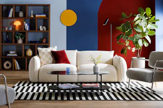 Wohnzimmer in blau-roter Farbgestaltung, inspiriert von den Designepochen Bauhaus und Mid-Century, im Zentrum ein helles Sofa, dazu graue Sessel mit Chromgestell und ein Couchtisch aus Rauchglas, dahinter ein großes Regal mit dunklem Walnussfurnier; dekoriert ist das Setting mit grafisch gemusterten Textilien und Accessoires in geometrischen Formen.