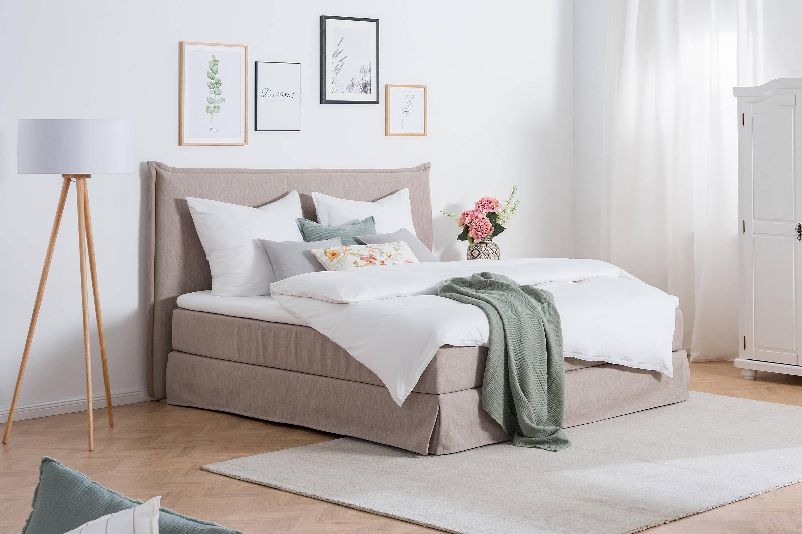Ländlich eingerichtetes Schlafzimmer, als Blickpunkt Boxspringbett Avellino mit Husse, gemütliche Decken, Kissen und Blumen, daneben ein großer weißer Kleiderschrank mit verzierten Drehtüren.