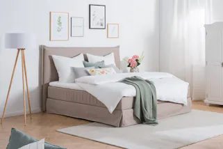 Ländlich eingerichtetes Schlafzimmer, als Blickpunkt Boxspringbett Avellino mit Husse, gemütliche Decken, Kissen und Blumen, daneben ein großer weißer Kleiderschrank mit verzierten Drehtüren.