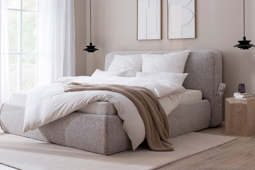 Erholsames Schlafzimmer in warmen Grau-, Beige- und Holztönen mit einem grauen Polsterbett, einem dunkelgrauen Sessel, passender Bettwäsche und einem Sideboard aus hellem Holz.